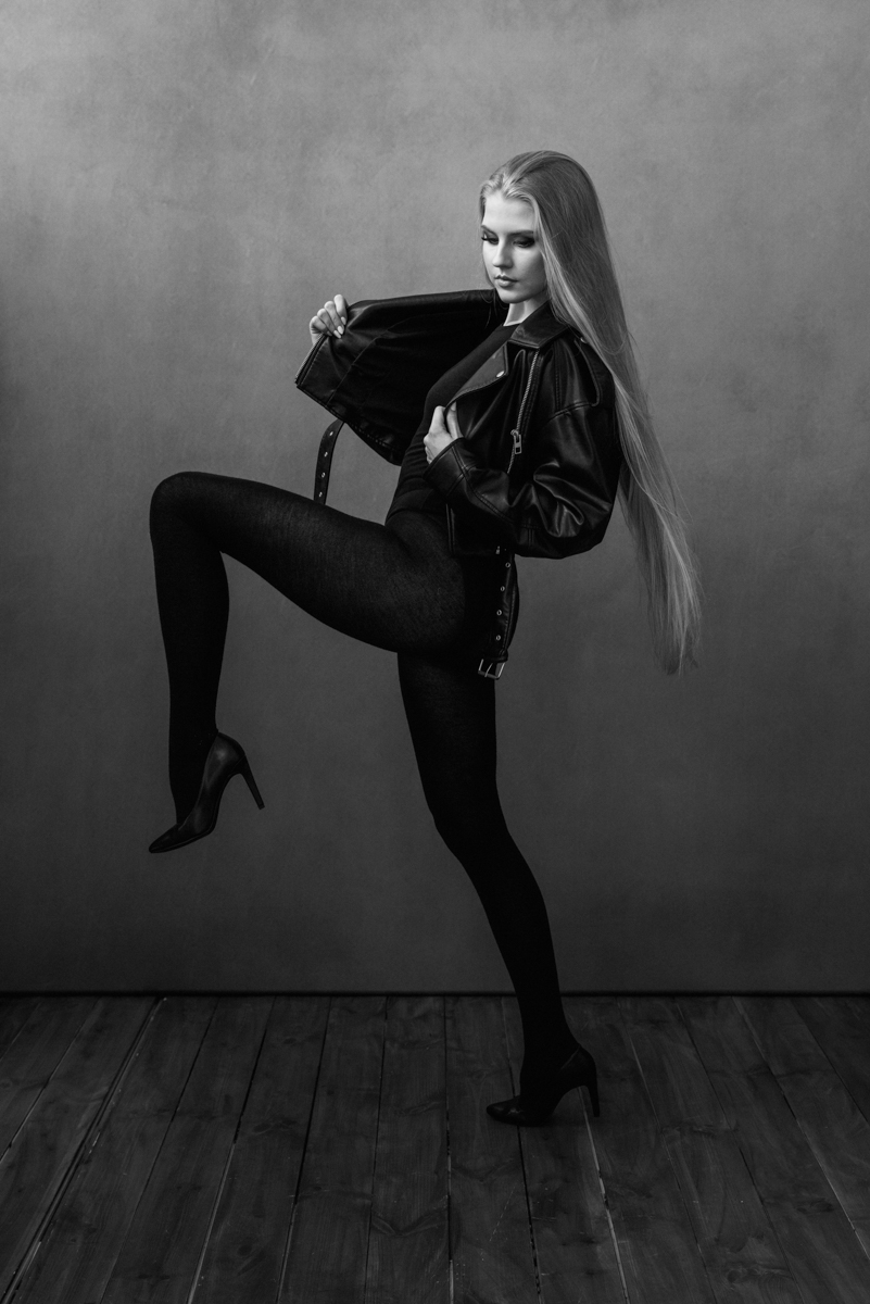 Mustvalge stuudioportree naisest dünaamilises poosis: keha profiilis, tõstetud jalg, pikk juus, nahktagi ja kontsakingad minimalistlikul taustal