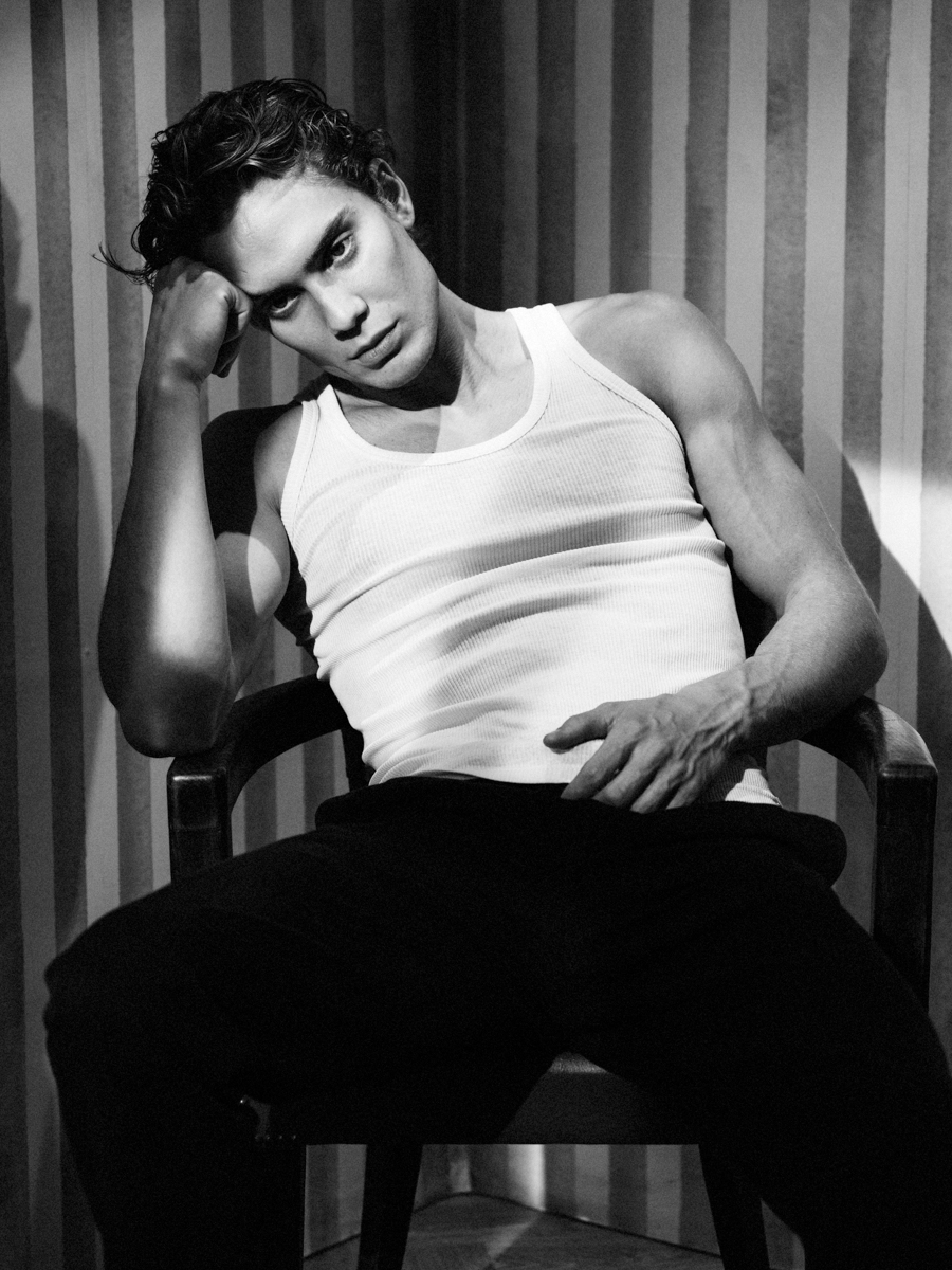 Mustvalge portree mehest, kes istub toolil triibulise seina taustal, kandes valget maikasärki ja vaadates mõtliku ilmega kaamerasse.